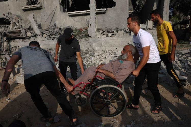 ثلاثة رجال يساعدون رجلا في كرسي متحرك على التنقل وسط الركام والدمار في جنوب قطاع غزة في 22 أكتوبر 2023.  © 2023 سعيد خطيب/أ ف ب